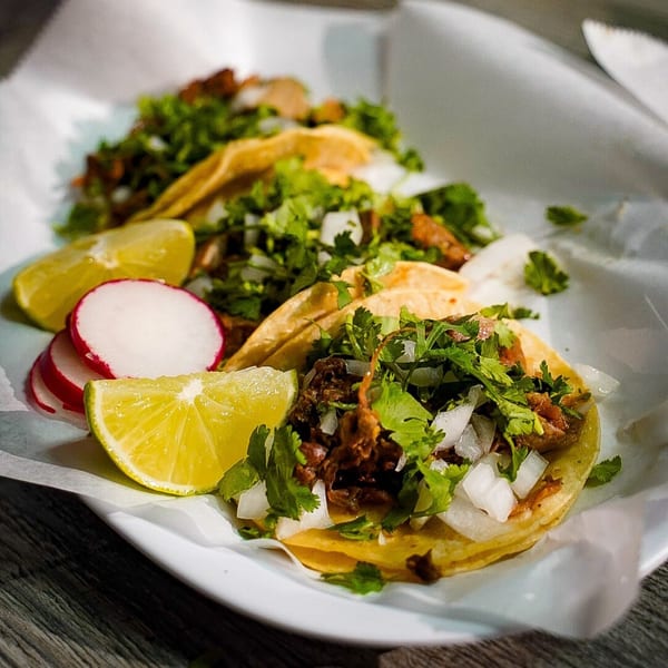 Three Las Salsas Taqueria tacos with meat, cilantro, onions, lime, and radishes on a white plate.