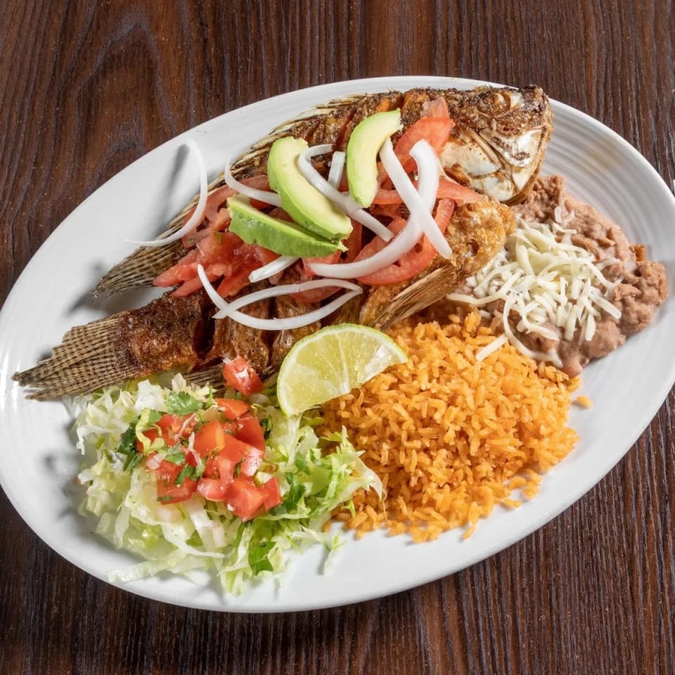 Whole fried fish with avocado, onions, tomatoes, rice, beans, and a lime wedge on a white plate.