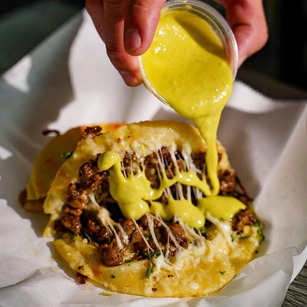 Hand pouring yellow sauce from a plastic cup onto cheesy Las Salsas Taqueria tacos.