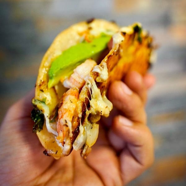 A hand holds a shrimp taco from Las Salsas Taqueria, filled with melted cheese and avocado.
