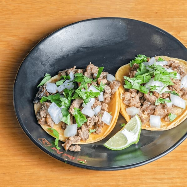 Two Las Salsas Taqueria tacos with meat, onions, cilantro, and a lime wedge on a black plate.