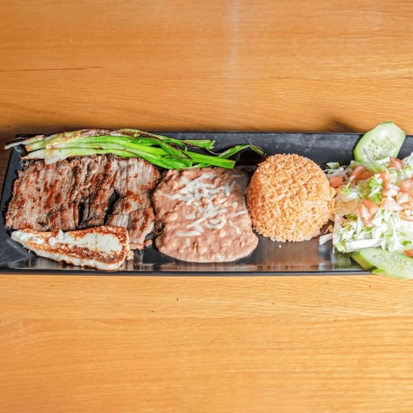 Grilled steak, refried beans, rice, grilled green onions, and salad with cheese on a black plate.