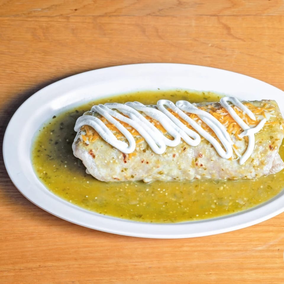 A large burrito covered in green salsa, sour cream, and shredded cheese on a white plate.
